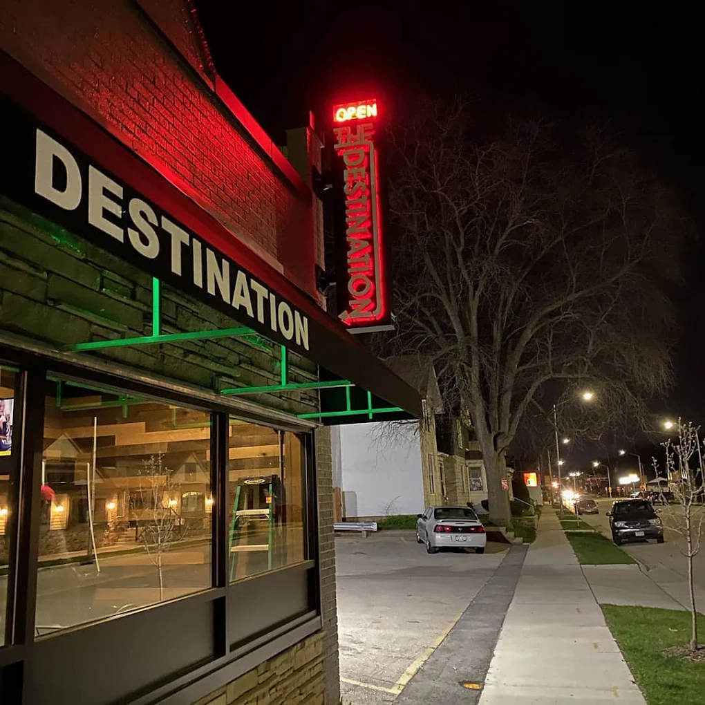 The Destination storefront at night with the red vertical neon sign lit up on E Main Street
